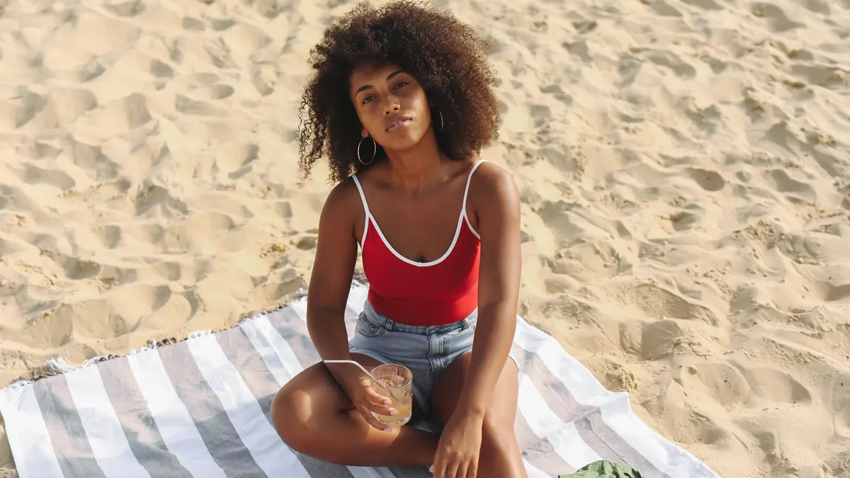 Young woman relaxing on a striped beach towel on the sand with a drink during her annual leave