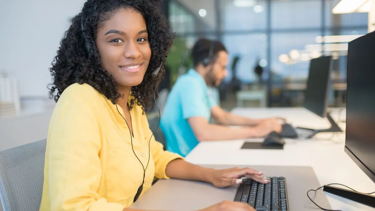 Smiling young Black woman in a yellow blouse wearing a headset at a computer in a busy call centre