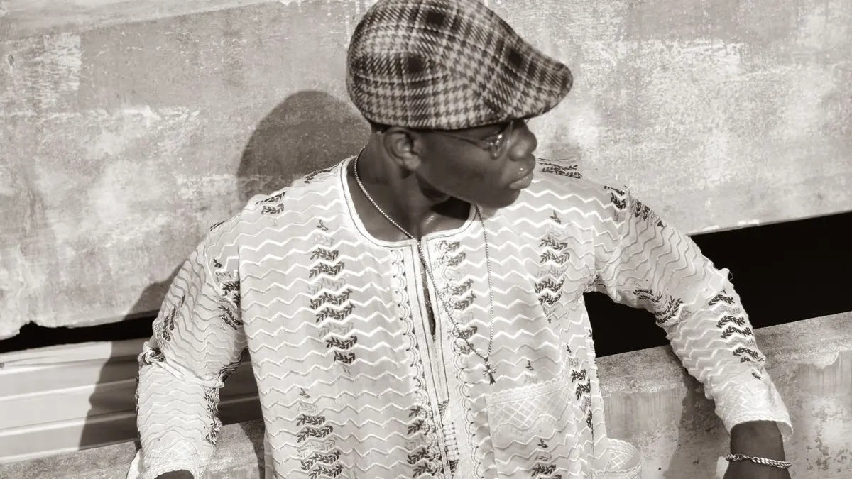 Black-and-white photo of an African man in a cap and patterned shirt carrying a briefcase