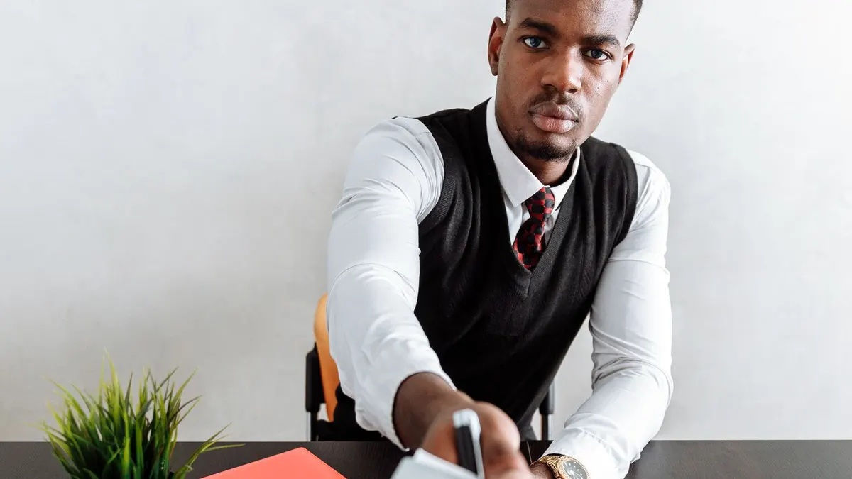 Young man in a white shirt and dark waistcoat handing a sheet of paper across an office desk