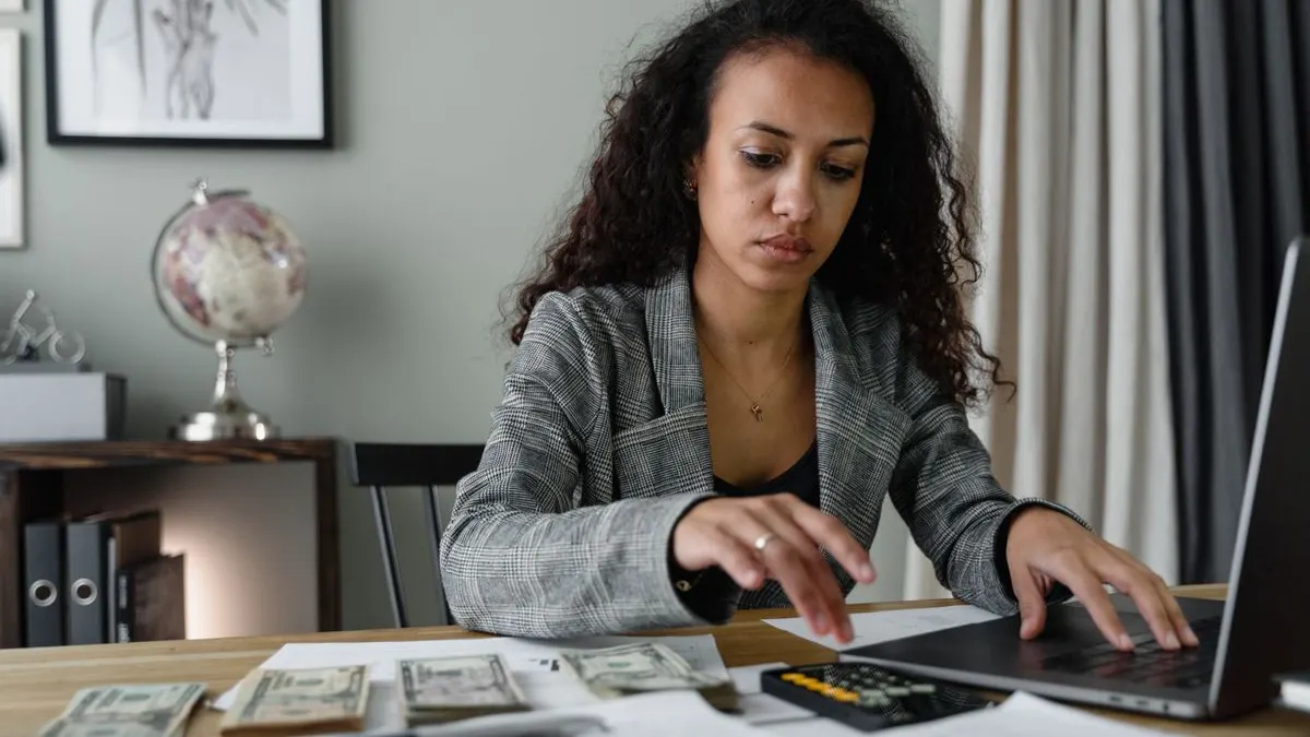 Young Black woman in a plaid blazer working on a laptop and phone at a desk covered with financial papers
