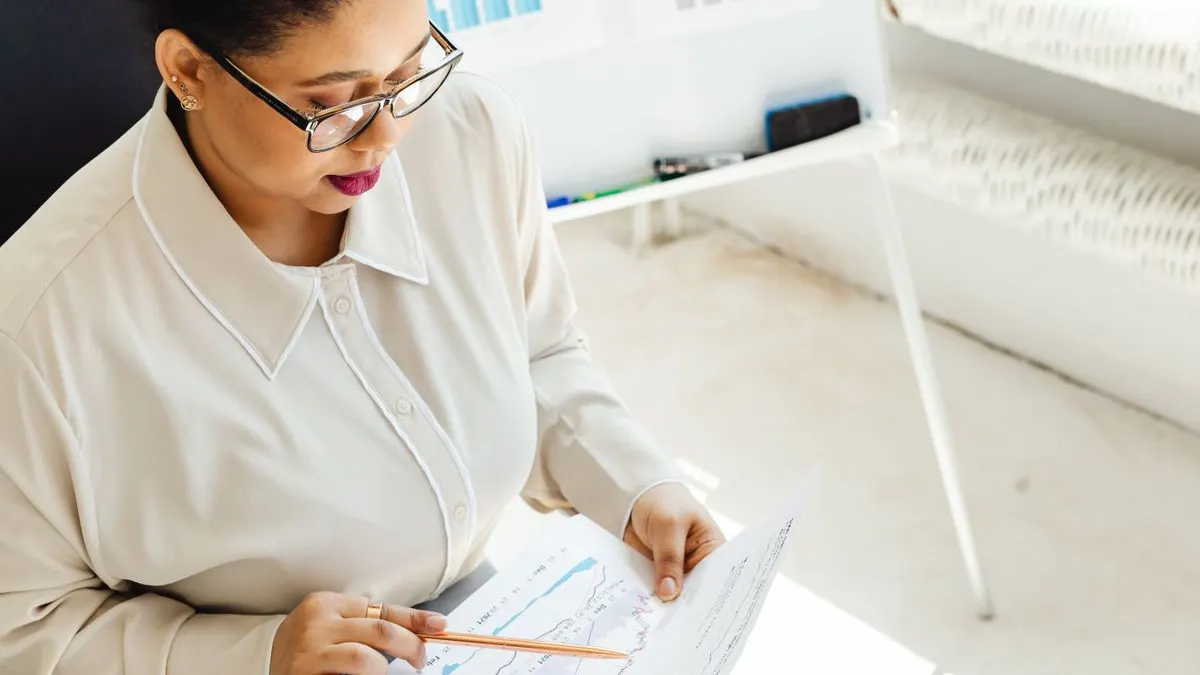 Black woman in glasses pointing a pencil at a printed line graph while reviewing data at her desk