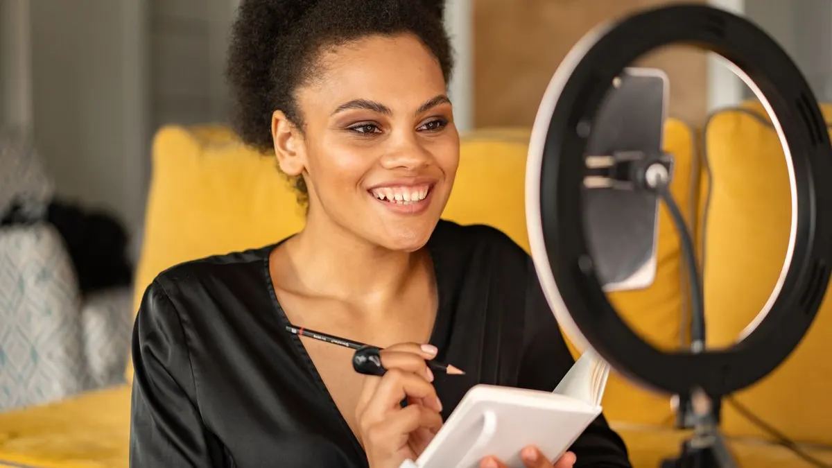 Smiling young Black woman with a notebook and pencil sitting in front of a ring light and phone while recording content