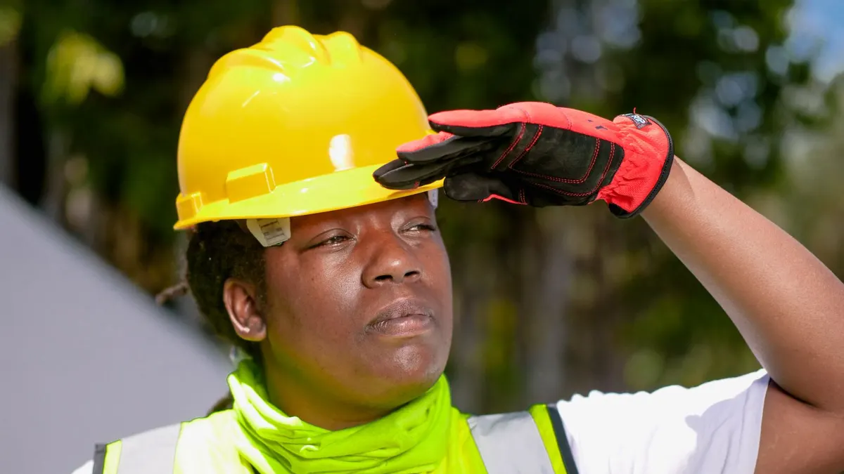 Black female engineer in a yellow hard hat and high-visibility vest shading her eyes on site