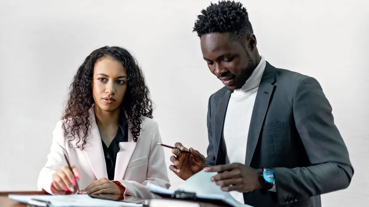 Young man in a grey suit pointing at a document as a female colleague reviews it at an office desk