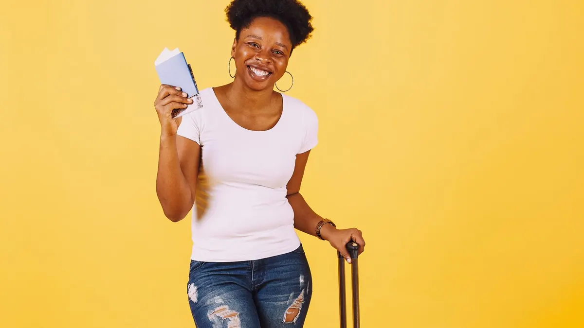 Smiling young Black woman holding a passport and standing beside a packed suitcase, ready to travel