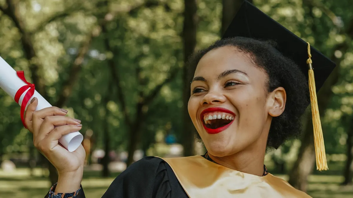 Smiling young Black woman in a graduation gown and cap holding her degree certificate