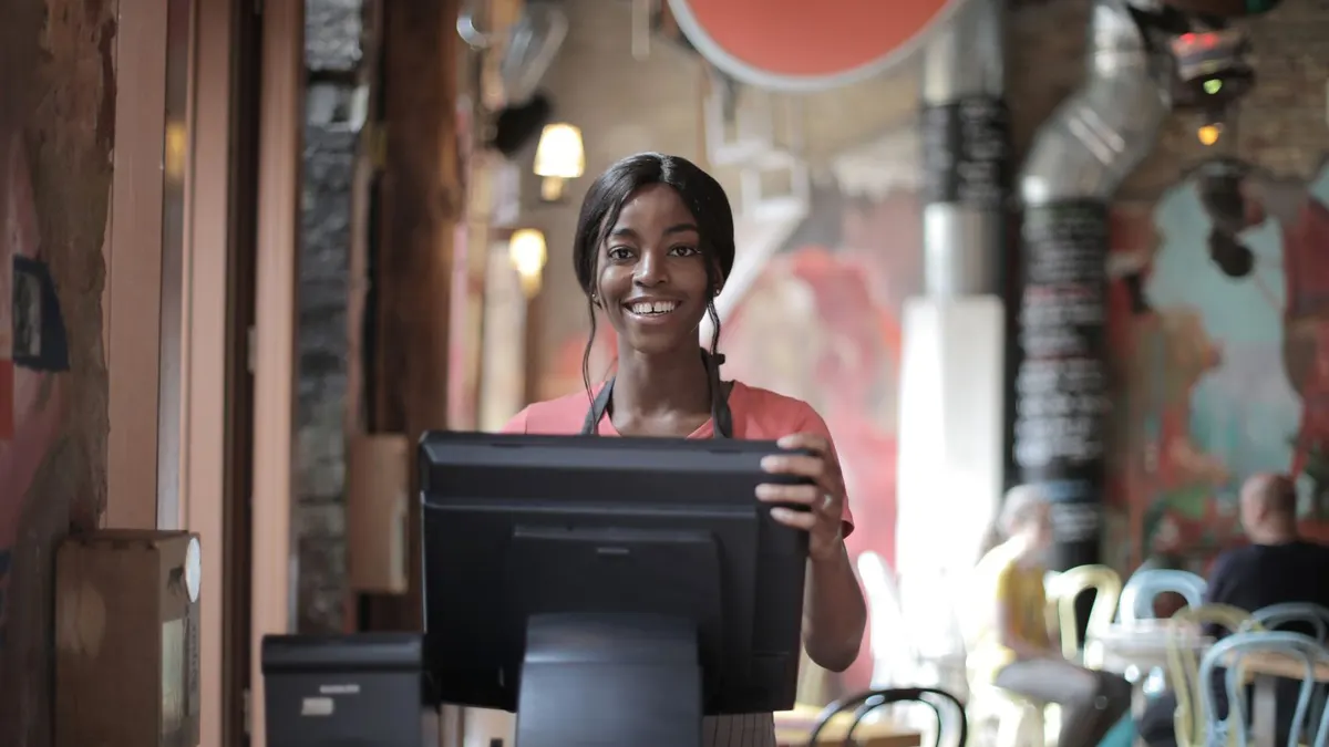 Smiling young Black waitress standing behind the counter of a busy restaurant