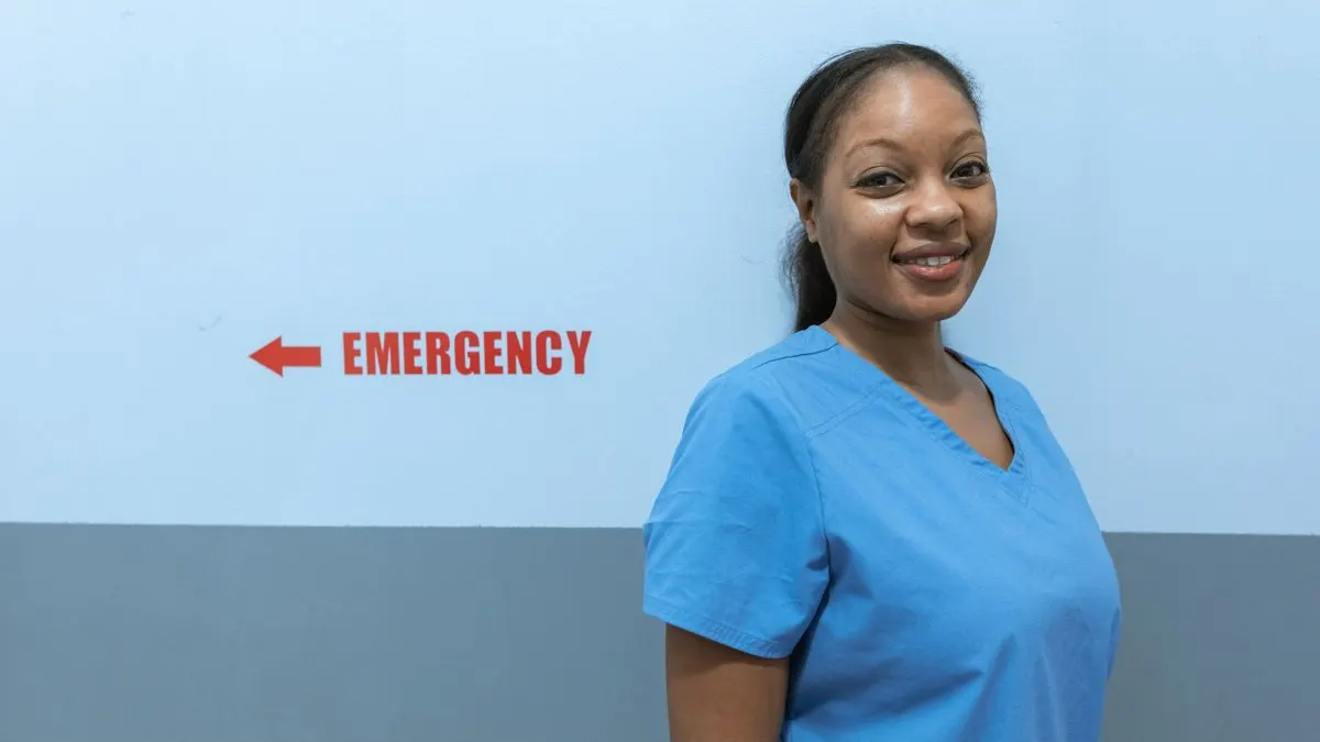 Smiling young Black nurse in blue scrubs standing in a hospital corridor next to an emergency sign