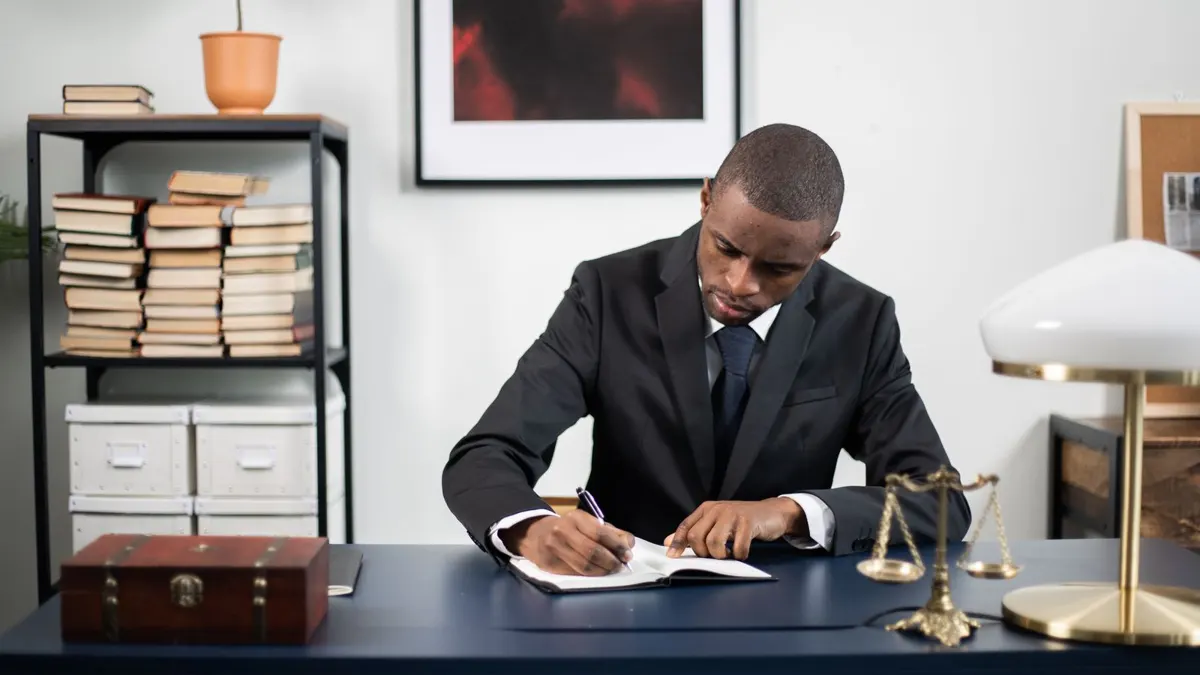 Man in a dark suit writing notes at an office desk with a scale of justice beside him