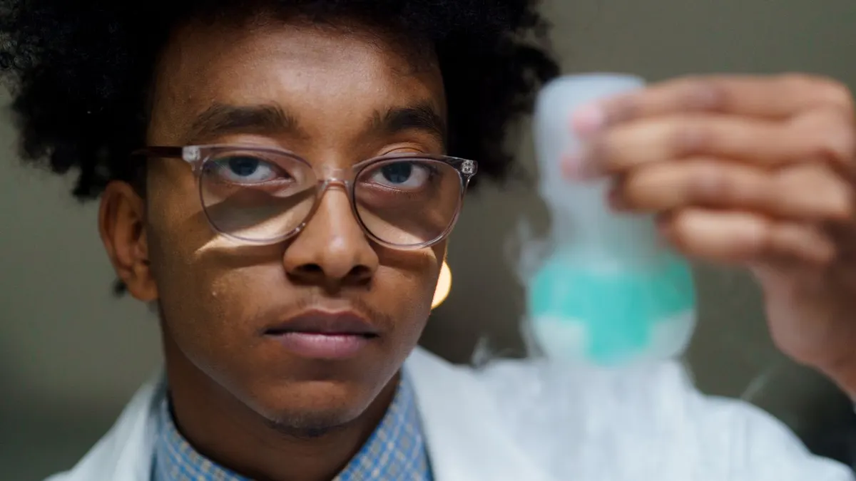 Young Black student in a lab coat and glasses holding up a flask of blue liquid