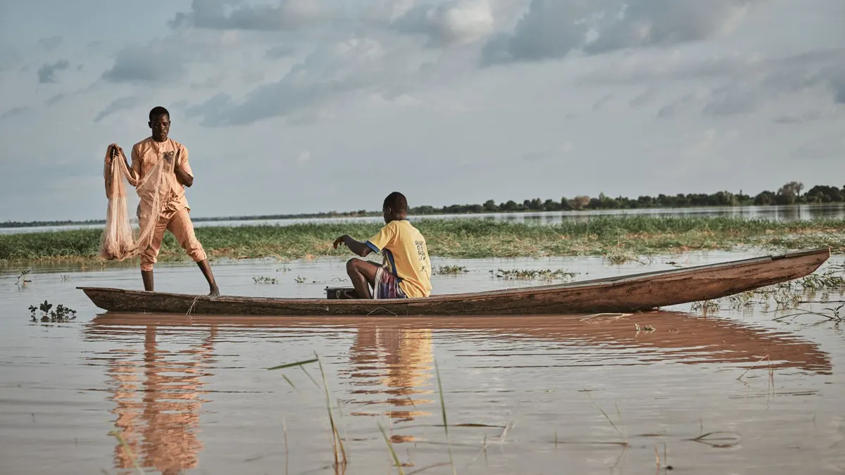 Two African fishermen in a wooden boat on calm lake water, one casting a net