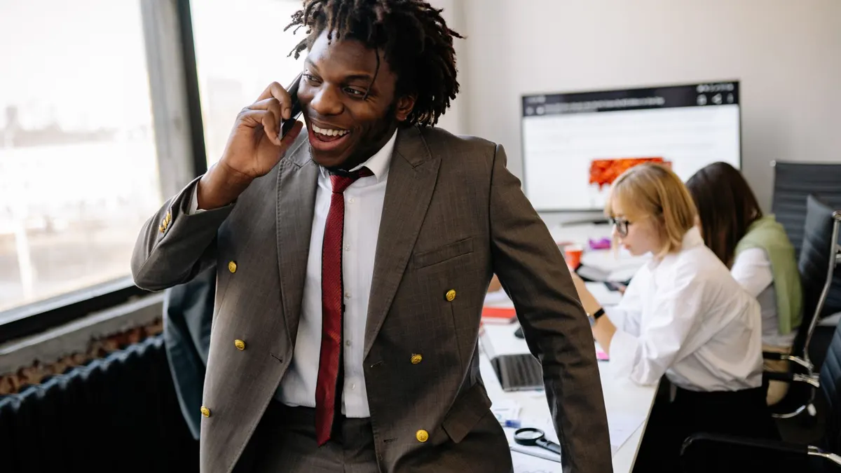 Smiling young Black man in a suit taking a phone call in a busy office
