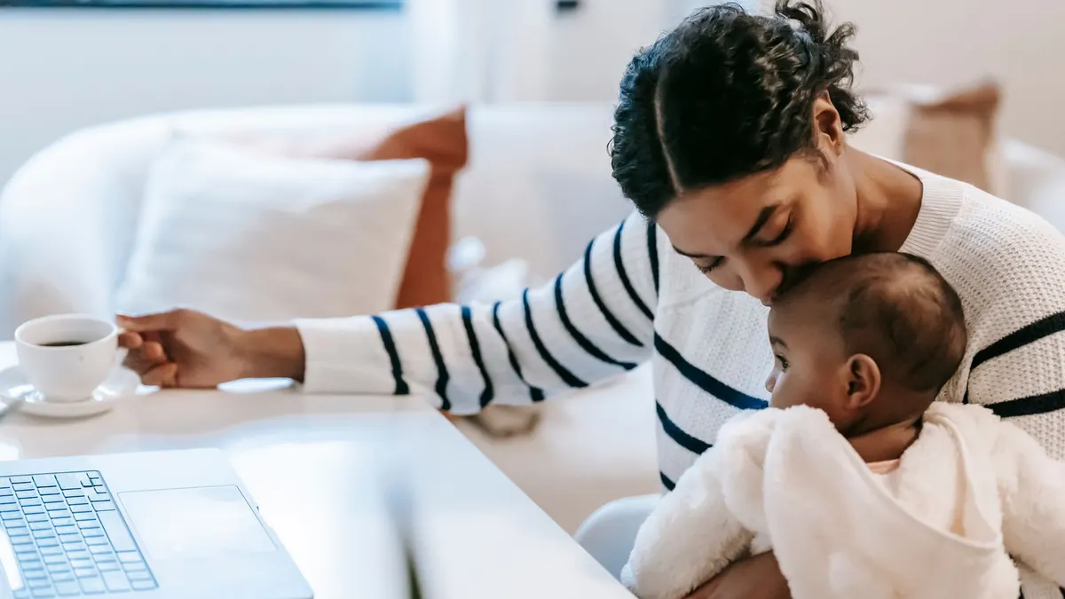 Working mother kissing her baby at home next to her laptop and a cup of coffee