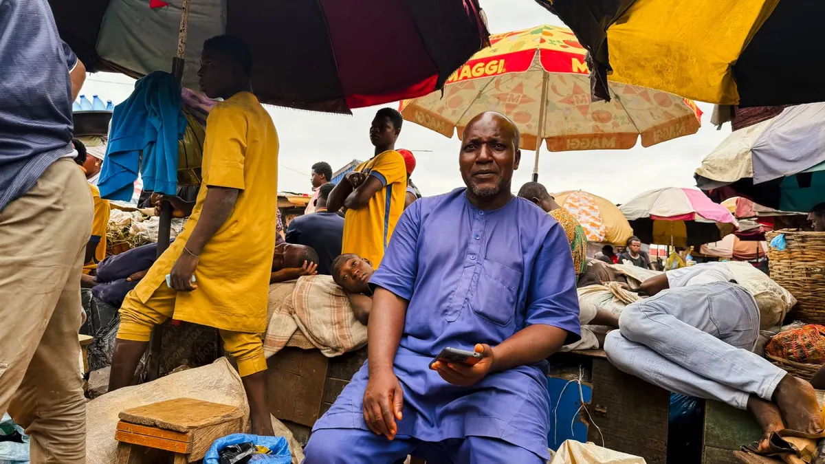 African trader in a blue outfit sitting at his stall in a busy open-air market under umbrellas