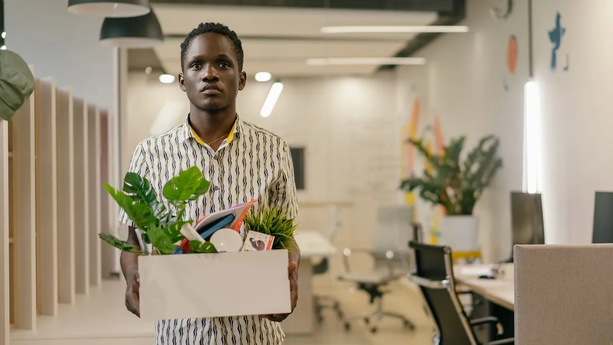 Young man in a patterned shirt standing in an empty office holding a white box with a potted plant and his belongings