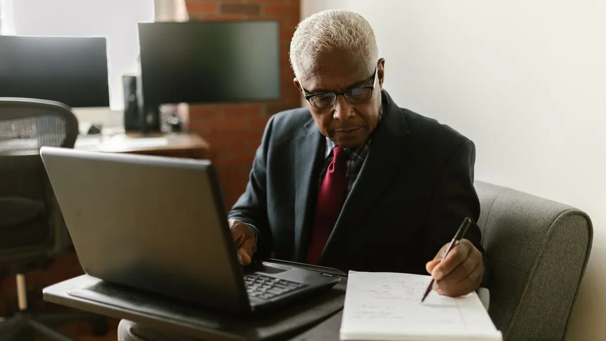 Older man in a dark suit writing notes beside a laptop at an office desk