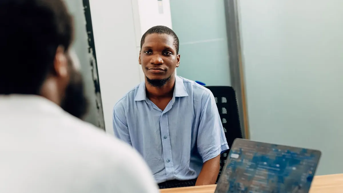 Smiling young Black man in a blue shirt facing an interviewer across a desk with a laptop