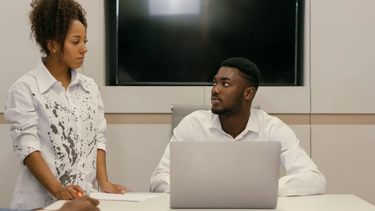 Woman standing at a desk speaking to a seated young man in a white shirt during a serious meeting in an office