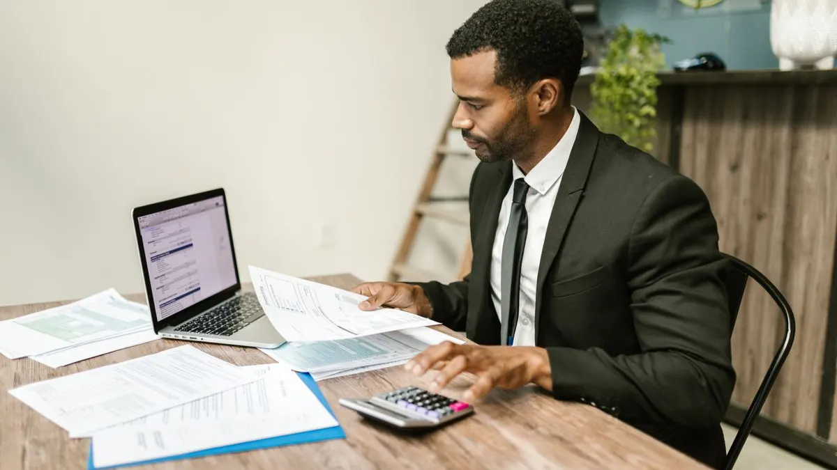 Black man in a dark suit working through papers with a calculator and laptop at a wooden desk