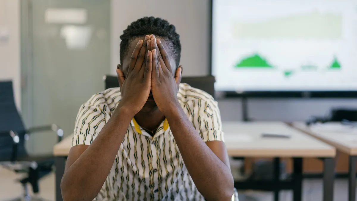Distressed young man in a patterned shirt sitting at an office desk with his face buried in his hands