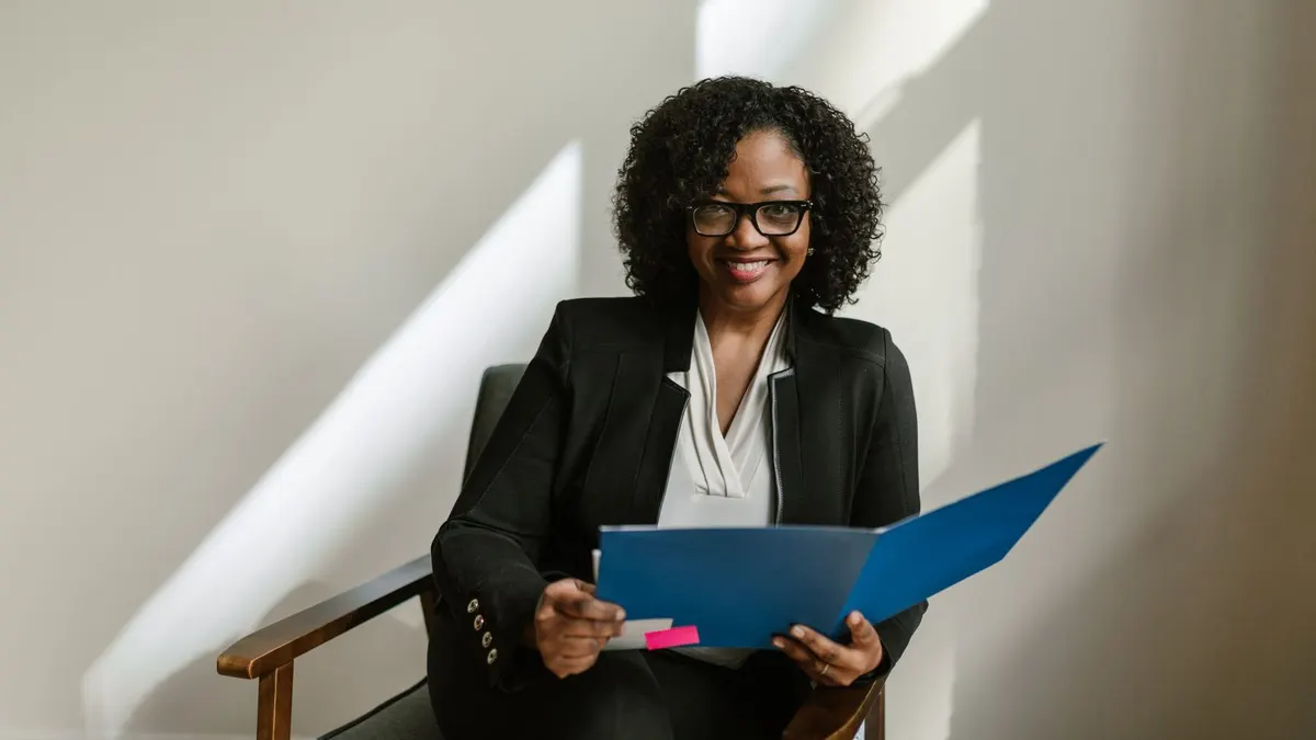 Smiling Black woman in a black blazer holding a blue folder while seated for an interview
