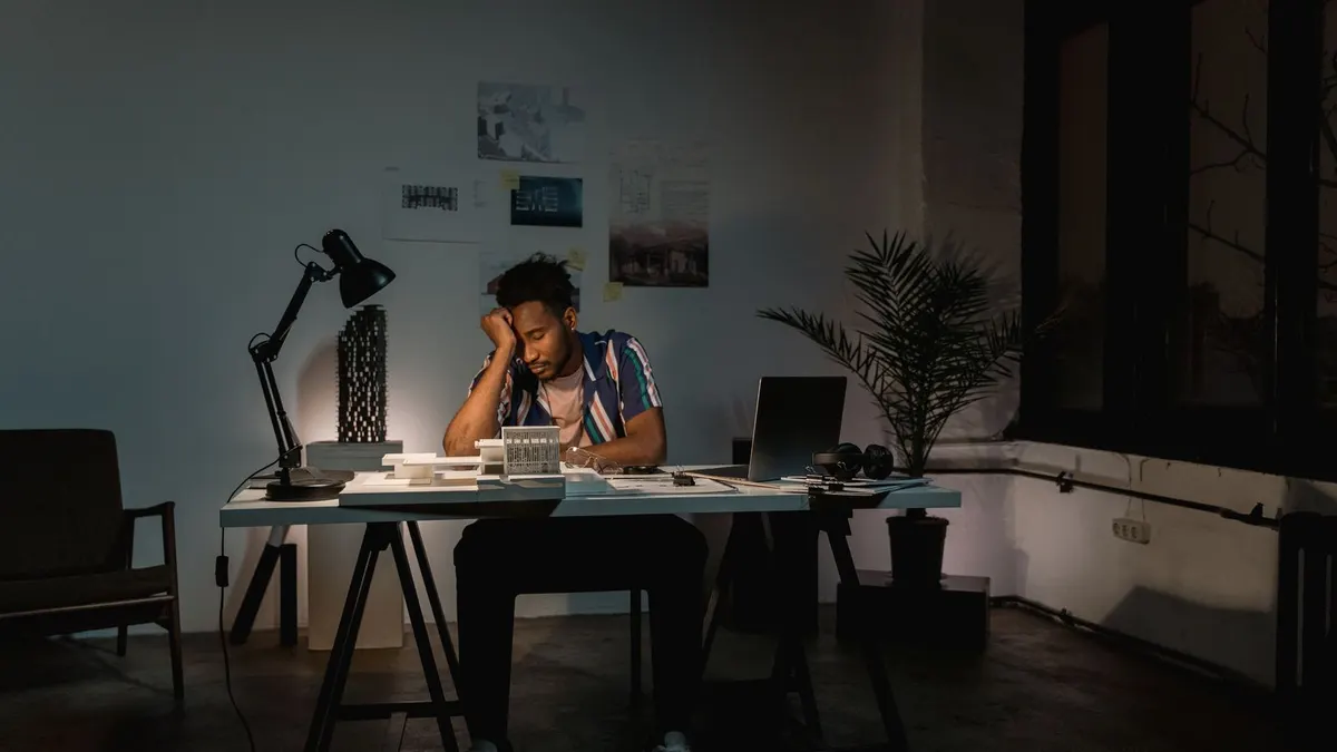 Tired young man resting his head on his hand at a desk in a dim office after hours